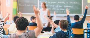 In einem Klassenraum sitzen Schüler an ihren Tischen und melden sich, vorne steht eine Lehrerin.