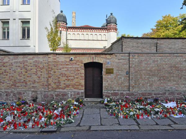 Vor einer gemauerten Wand mit einer Holztür stehen Kerzen und Blumen auf dem Bürgersteig.
