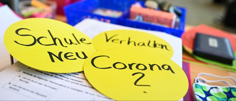 Schüler einer Abschlussklasse sitzen in der Achtalschule in Baden-Württemberg im Klassenzimmer, während auf dem Lehrerpult drei kreisrunde Papierschilder mit der Aufschrift Schule neu, Verhalten und Corona liegen.