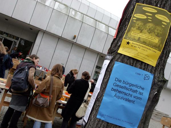 An einem Baum vor einem Unigebäude und Büchertischen hängen zwei Plakate.
