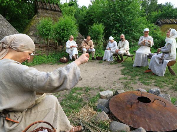 Menschen in historischen Kostümen aus Leinen sitzen im Kreis auf einem mittelalterlichen Dorfplatz.