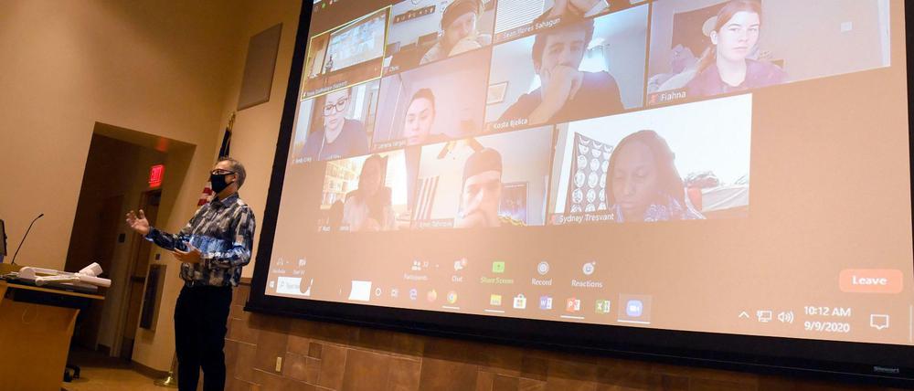 Ein Professor steht in einem Hörsaal, hinter ihm sind auf einer Leinwand Studierende zu sehen, die seine Vorlesung online verfolgen.