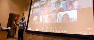 Ein Professor steht in einem Hörsaal, hinter ihm sind auf einer Leinwand Studierende zu sehen, die seine Vorlesung online verfolgen.