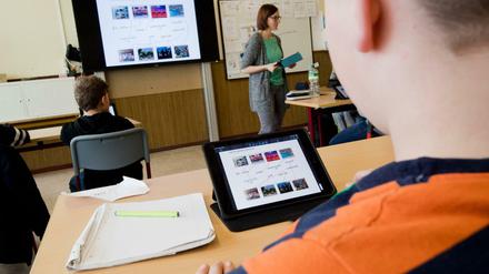 Schüler sitzen an in der Klasse vor IPads an ihren Tischen, vorne steht die Lehrerin vor einem großen Bildschirm.