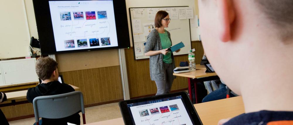 Schüler sitzen an in der Klasse vor IPads an ihren Tischen, vorne steht die Lehrerin vor einem großen Bildschirm.
