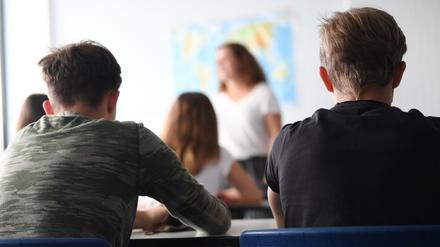 Zu sehen sind die Rücken und Hinterköpfe von Schüler:innen, vor denen einen Lehrerin steht.