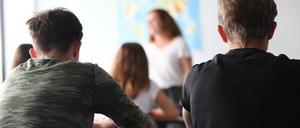 Zu sehen sind die Rücken und Hinterköpfe von Schüler:innen, vor denen einen Lehrerin steht.