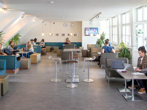 Studierende sitzen in einer Cafeteria auf Sesseln und Polsterbänken.