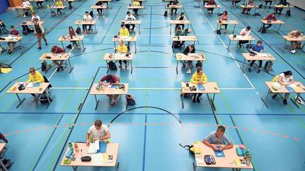 In einer Turnhalle sitzen Abiturienten und Abiturientinnen an Einzeltischen.