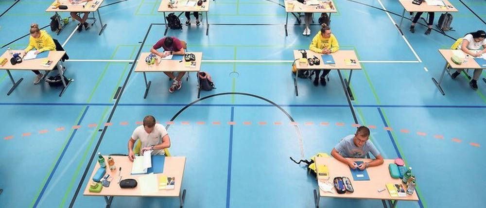 In einer Turnhalle sitzen Abiturienten und Abiturientinnen an Einzeltischen.