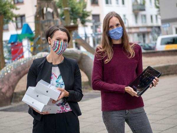 Zwei Frauen mit Alltagsmasken stehen mit Schachteln und Laptops auf einem Schulhof.