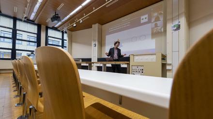 Ein Professor steht in einem leeren Hörsaal vor einer Leinwand, auf der Vorlesungsinhalte und weitere Teilnehmende zu sehen sind.