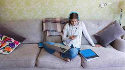 Eine junge Frau sitzt auf einem Sofa, hat ihr Laptop auf dem Schoß, ein Smartphone in der Hand und trägt Kopfhörer.