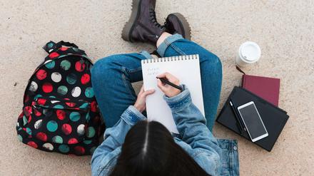 Eine Studentin sitzt im Schneidersitz am Boden und schreibt in einen Collegeblock.