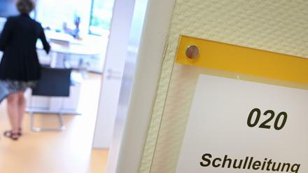 An einer geöffneten Tür steht "Schulleitung", im Raum ist eine Frau zu sehen, die sich an einen Tisch setzen will.