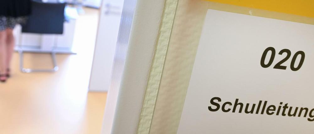 An einer geöffneten Tür steht "Schulleitung", im Raum ist eine Frau zu sehen, die sich an einen Tisch setzen will.