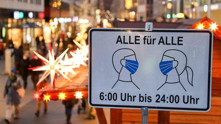 Passanten in Leipzig gehen hinter einem Schild entlang, das auf die Maskenpflicht in der Fußgängerzone hinweist.