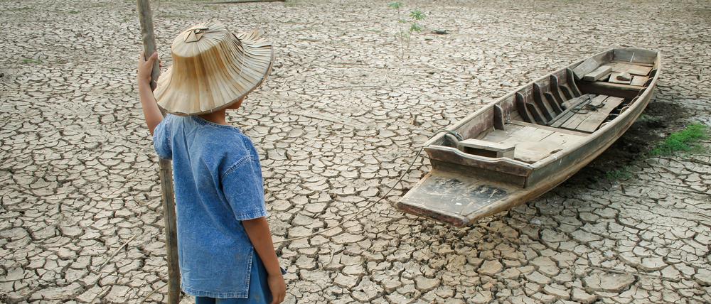 Symbolfoto: Ein Kind steht vor einem Holzboot, das auf ausgetrockeneter Erde liegt.