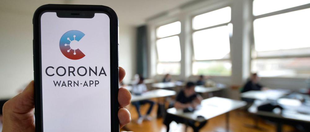 Hand hält Smartphone mit Corona-Warnapp vor Schulklasse.