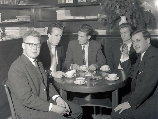 Fünf junge Männer in Anzug und Schlips sitzen vor Teetassen und Gläsern mit Alkohol an einem runden Tisch.