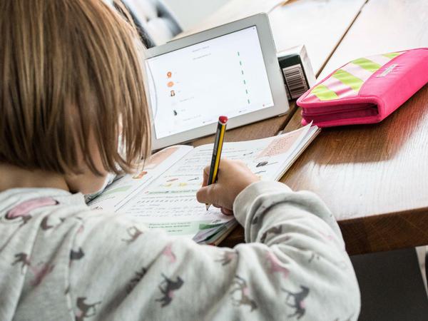 Eine Schülerin sitzt an ihrem Schreibtisch und schreibt in ein Schulbuch, vor ihr steht ein Tablet-Computer.
