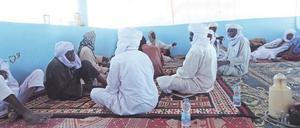 Männer in meist weißen Gewändern und mit turbanartigen Kopfbedeckungen sitzen in einem großen Raum auf Teppichen und reden miteinander.