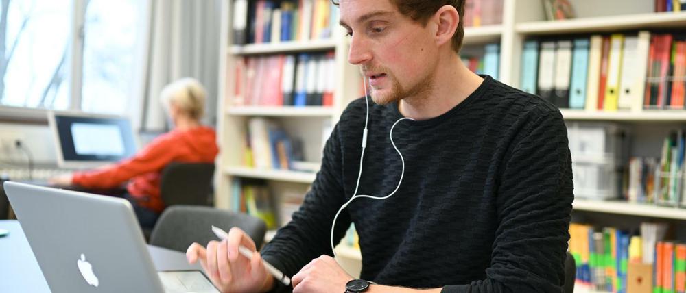 In einem Lehrerzimmer arbeitet ein Lehrer am Laptop, im Hintergrund sitzt eine Kollegin am Desktop.