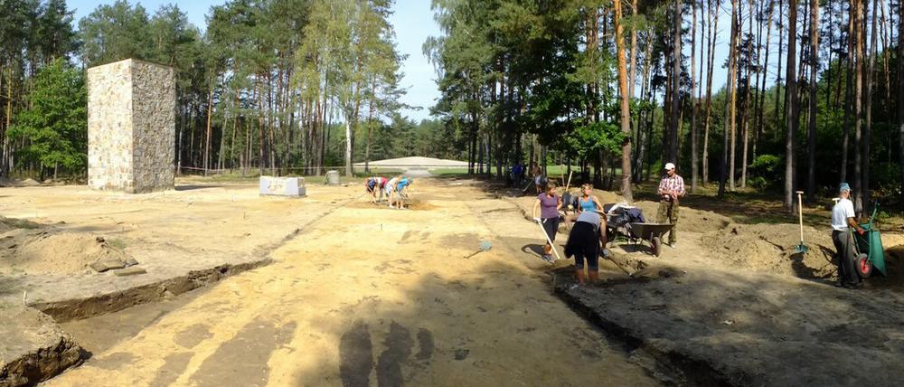 Archäolog:innen graben auf dem waldigen Gelände einer Gedenkstätte, im Hintergrund ist eine monumentale Gedenkstele zu sehen.