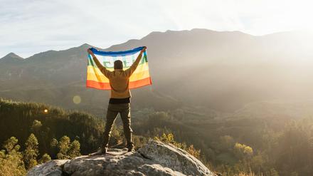 Der Mensch und die Natur. Ein Wanderer hält eine LGBTQ-Flagge hoch.