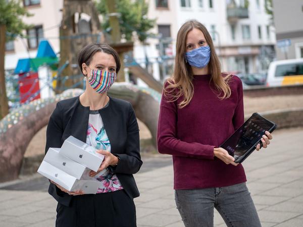 Zwei Frauen, die Mundschutz tragen, stehen mit Tablet-Computern im Arm auf einem Schulhof.