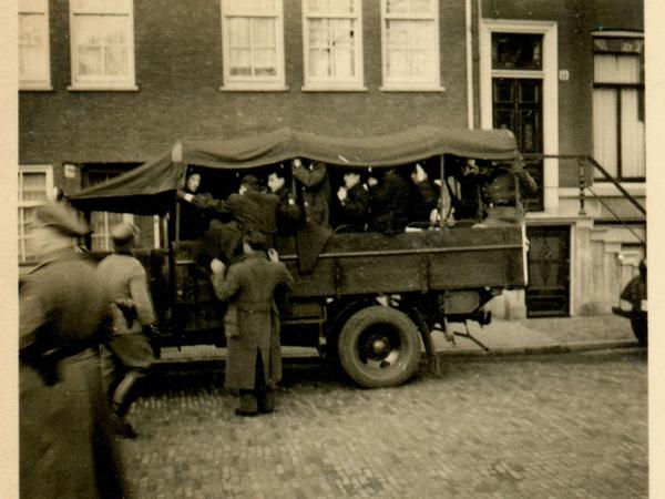 Auf einem Lastwagen sitzen Männern unter einer Plane, andere werden von Uniformierten gezwungen, ebenfalls einzusteigen.