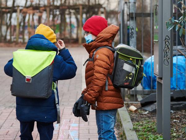Ein Junge setzt vor dem Schultor seine medizinisch Maske auf, ein anderer sieht ihm dabei zu.