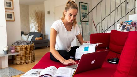 Eine Studentin sitzt mit ihrem Laptop und Lehrbüchern auf dem Sofa.