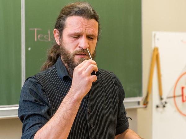 Ein Mann mit Bart und langem Zopf steht vor einer Tafel in einem Klassenzimmer und steckt sich ein Teststäbchen für einen Coronatest in die Nase.