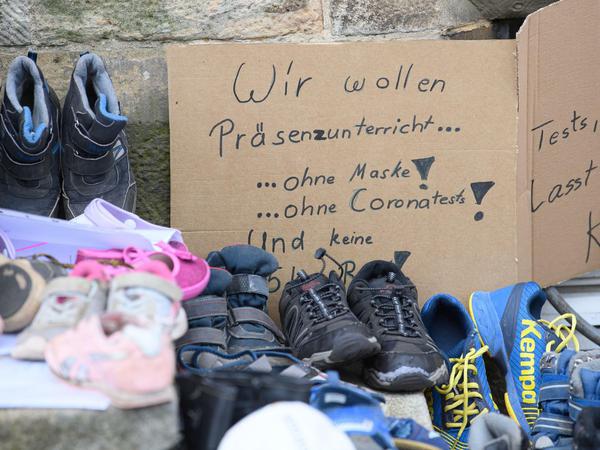 Hinter aufgereihten Kinderschuhen steht ein handgemaltes Plakat mit dem Text: Wir wollen Präsenzunterricht ... ohne Maske, ohne Coronatests!