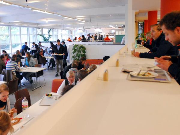 Studierende sitzen in einer Mensa, essen und unterhalten sich.