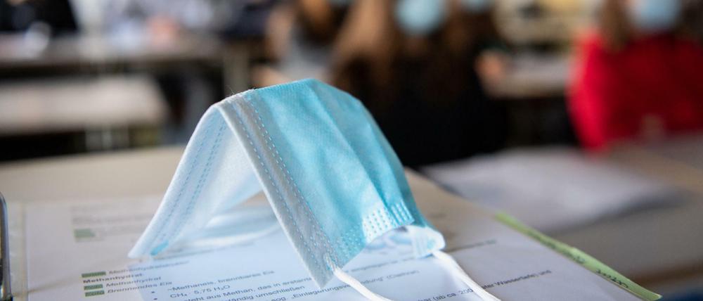 Eine Maske liegt im Unterricht auf Unterlagen, während im Hintergrund Schüler:innen mit Mund- und Nasenschutz zu sehen sind.