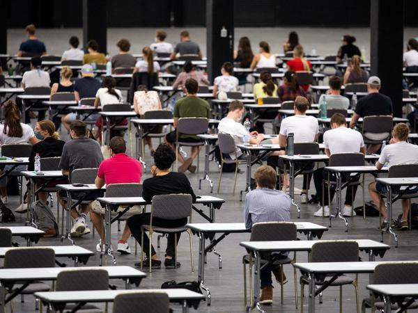 Studierende sitzen in einer Messehalle an Einzeltischen über ihren Prüfungsarbeiten.
