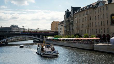 Menschen sitzen in den Außenbereichen von Cafés und Restaurants an der Spree, während ein Schiff vorbei fährt.