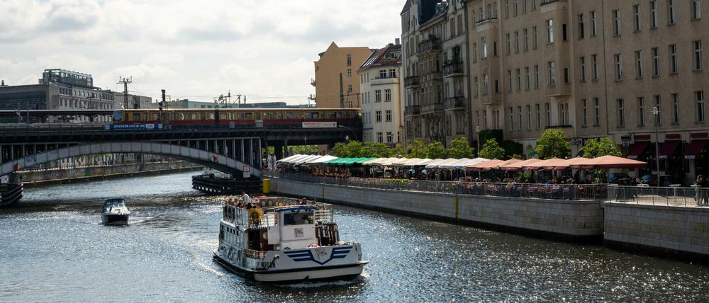 Menschen sitzen in den Außenbereichen von Cafés und Restaurants an der Spree, während ein Schiff vorbei fährt.