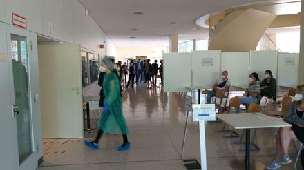 Eine junge Frau in einem grünen Schutzanzug geht durch das Foyer eines Unigebäudes, rechts davon sitzen Studierende in einem Wartebereich.