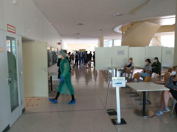 Eine junge Frau in einem grünen Schutzanzug geht durch das Foyer eines Unigebäudes, rechts davon sitzen Studierende in einem Wartebereich.