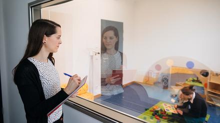 Eine junge Wissenschaftlerin macht sich bei der Beobachtung einer Kollegin, die in einer Kita-Situation mit einem Kind spielt, Notizen.