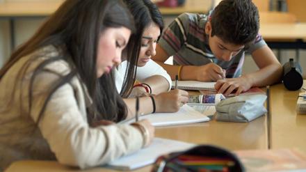 Jugendliche, zwei Schülerinnen und ein Schüler, beugen sich in einem Klassenraum über ihre Kollegblöcke und schreiben.
