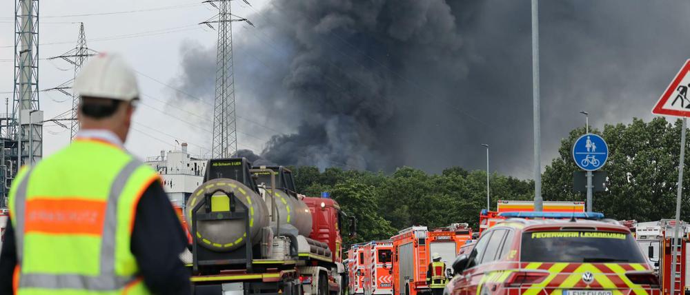 Einsatzfahrzeuge der Feuerwehr stehen unweit einer Zufahrt zum Chempark über dem eine dunkle Rauchwolke aufsteigt. 