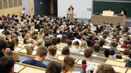 Studierende sitzen in einem Hörsaal, vorne spricht ein Dozent, an einem anderen Pult sind studentische Hilfskräfte zu sehen.