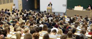 Studierende sitzen in einem Hörsaal, vorne spricht ein Dozent, an einem anderen Pult sind studentische Hilfskräfte zu sehen.