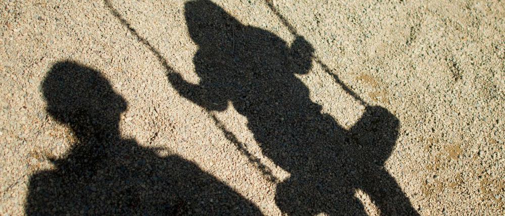 Ein Mann steht neben einem schaukelnden Kind, die Schatten zeichnen sich am Boden ab.
