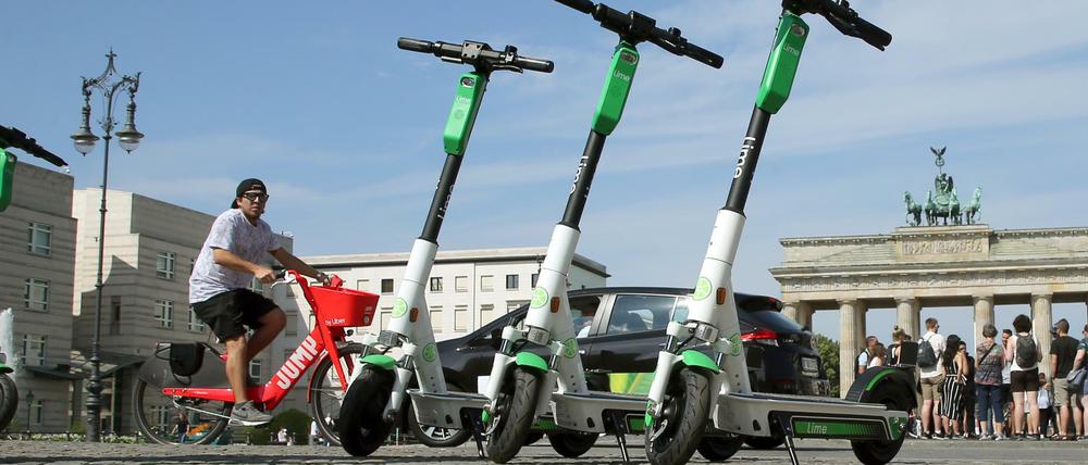 E-Tretroller der US-amerikanischen Firma Lime stehen vor dem Brandenburger Tor, während im Hintergrund ein Mann auf einem E-Bike des Anbieters Uber vorbeifährt. (Archivbild)