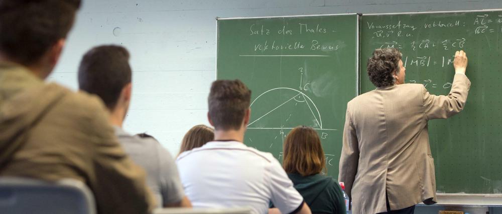 Ein Mathematiklehrer steht mit dem Rücken zur Klasse und schreibt mit Kreide Mathematikaufgaben an eine Tafel.